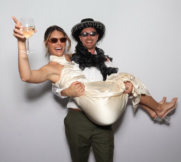 A man wearing a hat and sunglasses carrying a woman in a satin dress